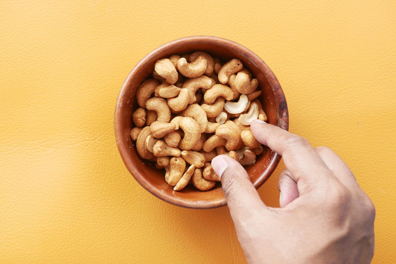 A close-up of a hand reaching for cashews in a wooden bowl on a vibrant yellow background.